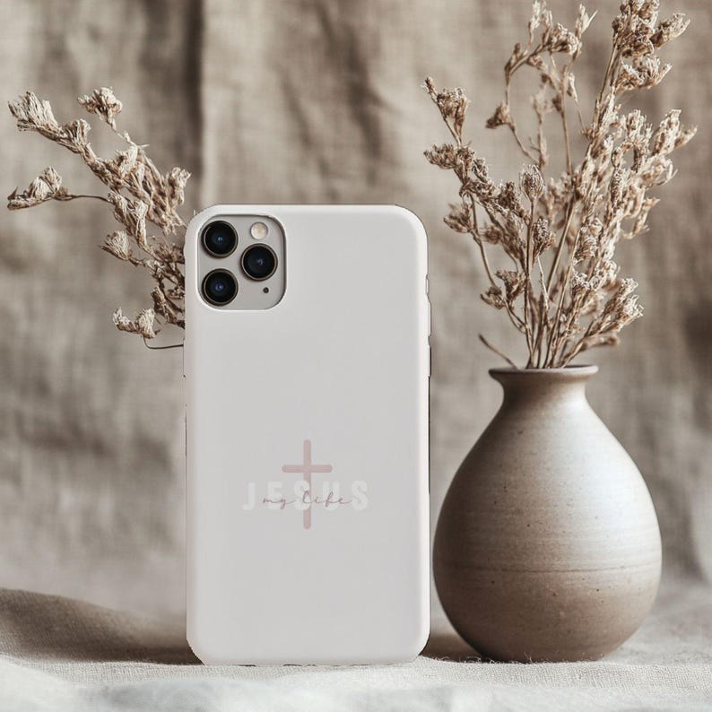May include: A white phone case with the words "Jesus my life" and a cross graphic in a soft pink color. The phone case is next to a vase with dried flowers.