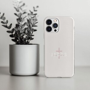 May include: A white phone case with a pink cross and the words "Jesus my life". The case is next to a potted plant with green leaves.