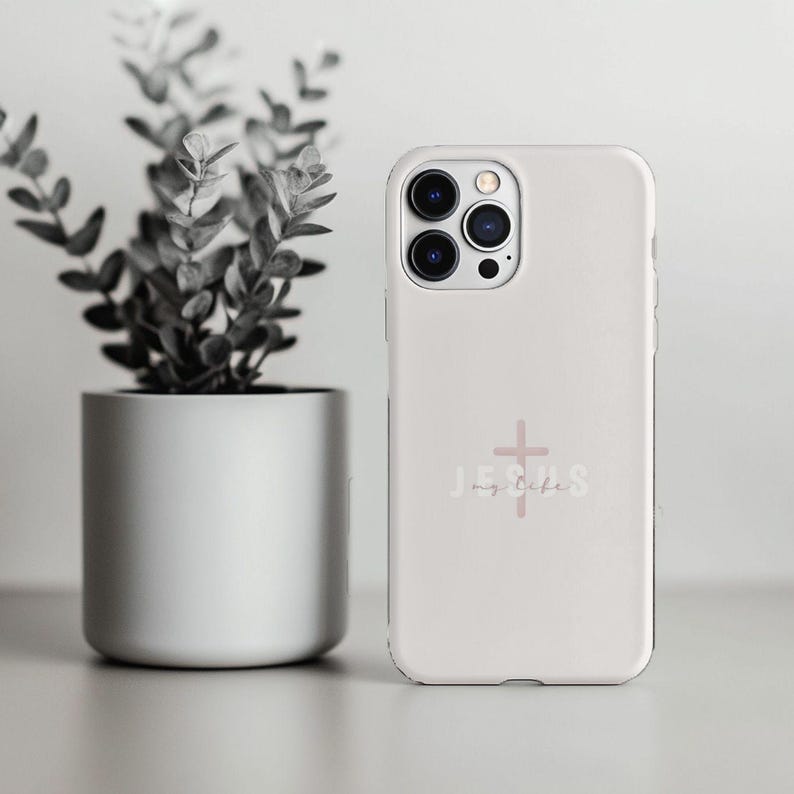 May include: A white phone case with a pink cross and the words "Jesus my life". The case is next to a potted plant with green leaves.