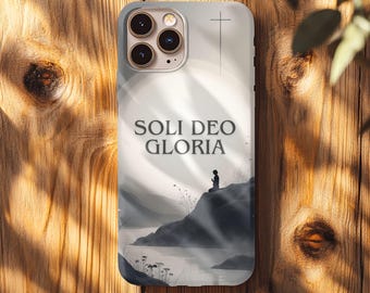 May include: A phone case with a serene landscape design. The case features a silhouette of a person in prayer on a hillside, with the words "SOLI DEO GLORIA" and a cross. The background depicts a misty lake and mountains.