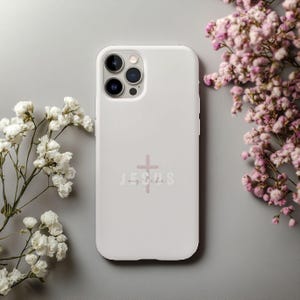 May include: A white phone case with the word "Jesus" and a cross in a soft pink color. The phone case is surrounded by white and pink flowers on a gray background.