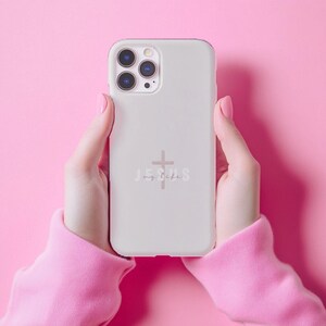 May include: A light gray phone case with a cross and the words "Jesus my life" in a script font. The phone case is held against a pink background. The case has a camera module with three lenses.