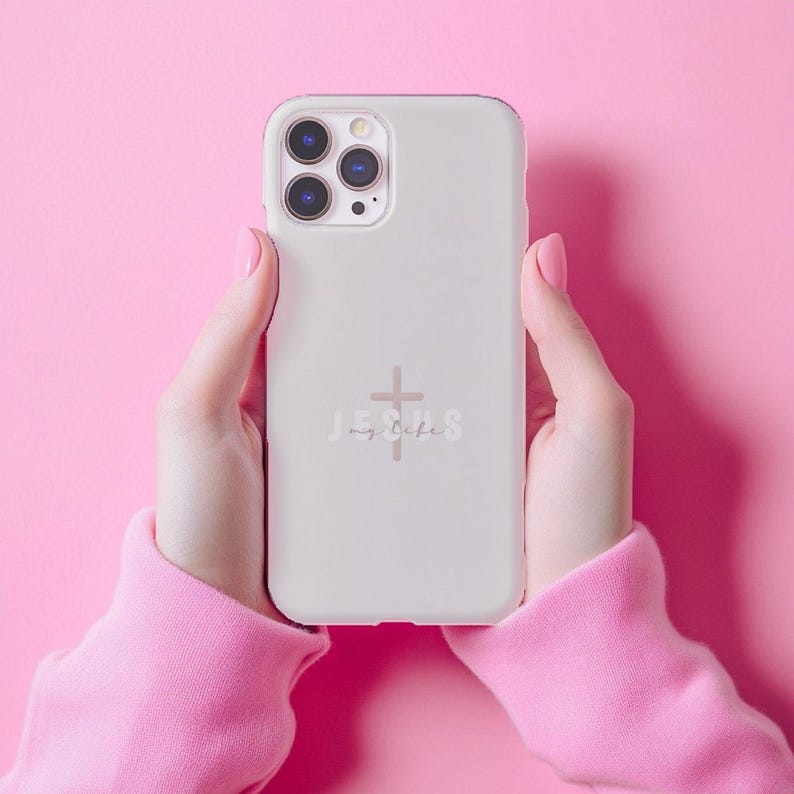 May include: A light gray phone case with a cross and the words "Jesus my life" in a script font. The phone case is held against a pink background. The case has a camera module with three lenses.