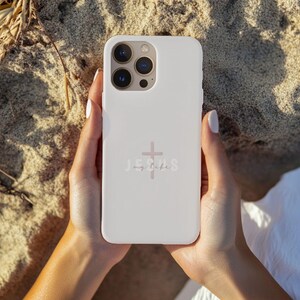 May include: A white phone case with a cross and the words "Jesus my life" in a light pink color. The phone case is held by a person with light skin, with the background of sand.