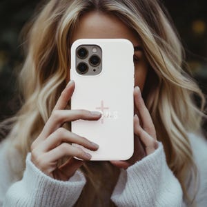 May include: A light pink phone case with a cross and the word "Jesus" printed on it. The case has a matte finish and a camera cutout. The phone case is held up in front of a person.