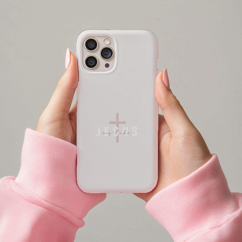 May include: A white phone case with the words "JESUS my life" and a cross graphic in a soft pink color. The phone case is held by hands wearing a pink sweatshirt.