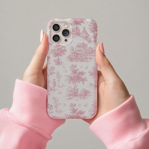 May include: A white and pink phone case featuring a toile pattern with trees, buildings, and figures. The case is held by a person wearing a pink sweatshirt. The case has a camera cutout and buttons.