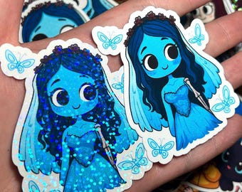 May include: A hand holds several stickers featuring a blue cartoon bride with dark blue hair, a veil, and a blue dress. The stickers have a holographic effect and butterfly accents. Other stickers with similar designs are visible.