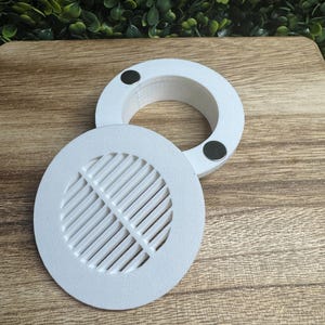 Puede incluir: Tapa de ventilación blanca de dos piezas con diseño circular. Una pieza presenta un patrón ranurado, mientras que la otra es un anillo liso. Ambas piezas tienen pequeños imanes negros. Las tapas de ventilación están sobre una superficie de madera.