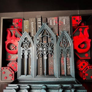 May include: A teal and black gothic-style bookshelf organizer with three arched sections. The organizer has a decorative design and is placed in front of several books. Red and black decorative boxes are on either side.
