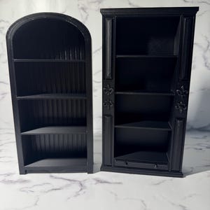 May include: Two black miniature bookshelves. One has an arched top and vertical slats on the back. The other has a rectangular shape with decorative molding, fleur-de-lis accents, and a small drawer. Both have multiple shelves.