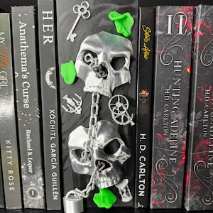 May include: A silver skull-themed bookmark with a chain, padlock, and key accents. The bookmark features two skulls, green accents, and is placed among several books on a shelf. The books have titles like "HER" and "Hunting Adeline".