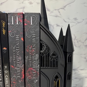 May include: Black gothic-style bookends with a cathedral design. The bookends are holding several books with the title "HAUNTING ADELINE" by H.D. Carlton. The bookends have intricate window details and a tall spire.
