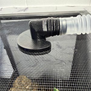 以下が含まれることがあります： 黒色の3Dプリントされた角度付きコネクタ。円形のベースと透明なフレキシブルチューブが取り付けられています。 コネクタは、おそらくエンクロージャー用の金属メッシュスクリーンに取り付けられています。