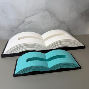 May include: Two open book-shaped stands, one larger in white and one smaller in turquoise, both featuring a black rectangular base. Each stand has a central horizontal slot cut into the top surface, designed to hold items in an upright position.