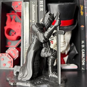 Könnte beinhalten: Eine schwarze, 3D-gedruckte Figur eines knienden Ritters, der ein Schwert hält. Der Ritter trägt einen Helm und eine Rüstung mit einem langen Umhang. Die Figur steht auf einem Regal mit Büchern und anderen dekorativen Gegenständen.