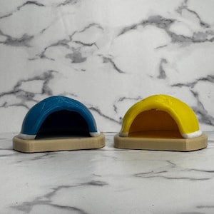May include: Two small, dome-shaped pet houses. One is blue with a white trim, and the other is yellow with a white trim. Both are set on a light brown base. The background is a marble pattern.