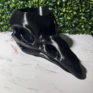 May include: A black, 3D-printed raven skull-shaped planter. The planter has a hollow interior for holding plants or other decorative items. The skull is detailed with eye sockets, a beak, and other anatomical features. The planter is sitting on a white marble surface.