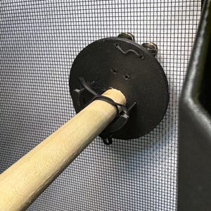 May include: A black, circular perch mount with a wooden dowel rod attached. The mount is secured to a mesh background with zip ties. The dowel rod is light brown. The background is a black mesh.