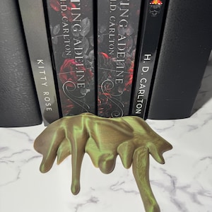 May include: A bronze-colored, abstract sculpture with a dripping design. The sculpture is in front of a row of black books with the title "Haunting Adeline" by H.D. Carlton. The sculpture has a metallic sheen and is on a white surface.