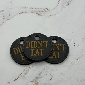 May include: Three circular black tags with a textured surface, each featuring the text DIDN'T EAT in raised gold lettering. Each tag has a small circular hole at the top for attachment, and the design is consistent across all three items.