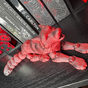 Puede incluir: Una figura de gato rojo articulada con una superficie texturizada detallada. El gato está tumbado con la boca abierta y la cola curvada. La figura está sobre una superficie negra, con libros visibles en el fondo.