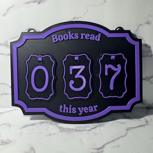 May include: A black and purple 3D-printed sign displaying "Books read this year" and the numbers "037". The sign has a decorative border and is designed to track the number of books read.