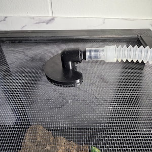 May include: A black plastic corner fitting with a clear, corrugated tube attached. The fitting is mounted on a black mesh screen, likely for a terrarium or similar enclosure. The background is a white tiled surface.