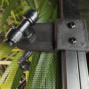 Puede incluir: Hardware de montaje de plástico negro con una junta giratoria y dos tornillos. El hardware está fijado a una pantalla de malla negra, con follaje verde visible en el fondo.