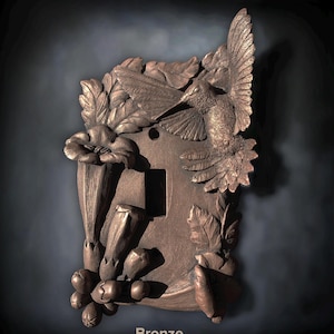 May include: A bronze-colored light switch plate featuring a three-dimensional relief sculpture of a hummingbird in flight, surrounded by detailed trumpet-shaped flowers and foliage. The word Bronze is printed at the bottom of the image.