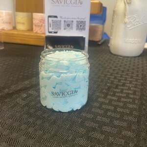 Puede incluir: Un tarro transparente lleno de un producto azul claro, similar a crema batida. El tarro tiene una etiqueta blanca con el texto "SAVICGIA LUXURY NATURAL PRODUCTS". Está sobre una superficie oscura con un patrón gris. Un letrero con el mismo logotipo está en el fondo.