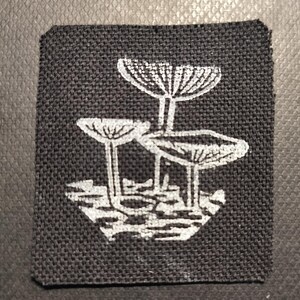 Può includere: Una toppa quadrata nera con un'illustrazione bianca di tre funghi. I funghi hanno cappelli e gambi dettagliati, su uno sfondo strutturato. La toppa è progettata per essere cucita o stirata sul tessuto.
