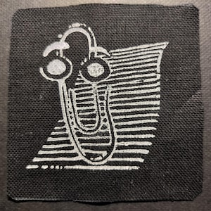 以下が含まれることがあります： 白の様式化されたデザインが施された正方形の黒いパッチ。デザインは、垂直のU字型要素とストライプの背景に接続されたヘッドフォンのペアを特徴としています。パッチは、おそらく衣類またはアクセサリー用です。