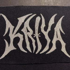 Könnte beinhalten: Ein schwarzer Stoffaufnäher mit dem weißen Wort "KRYA" in einer stilisierten, gotischen Schrift. Die Buchstaben haben scharfe, spitze Serifen, die einen kühnen und kantigen Eindruck erwecken. Der Aufnäher ist rechteckig mit abgerundeten Ecken.