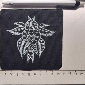May include: A black fabric patch with a white, stylized floral design. The patch is approximately 10 cm wide and 10 cm tall. A white pen rests above the patch. The patch is on a white surface with a cm ruler.
