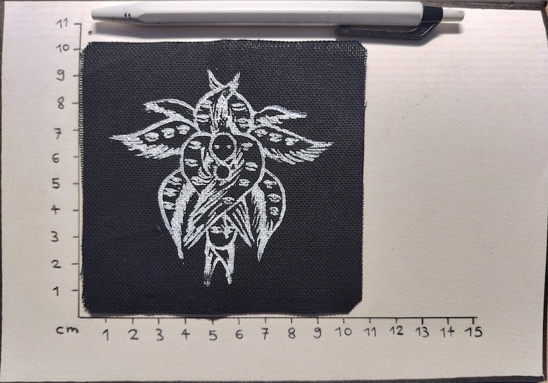 May include: A black fabric patch with a white, stylized floral design. The patch is approximately 10 cm wide and 10 cm tall. A white pen rests above the patch. The patch is on a white surface with a cm ruler.