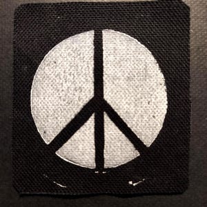 May include: A square, black fabric patch featuring a white peace symbol. The peace symbol is a circle with three lines inside, representing peace. The patch is likely for clothing or accessories.