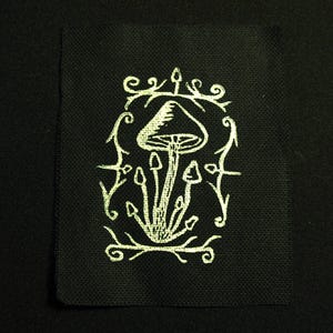 Könnte beinhalten: Schwarzer quadratischer Aufnäher mit einem weißen, gestickten Pilzdesign. Das Design zeigt einen großen Pilz mit kleineren darunter, umrahmt von einem dekorativen Rand. Der Aufnäher ist aus einem strukturierten Stoff gefertigt.