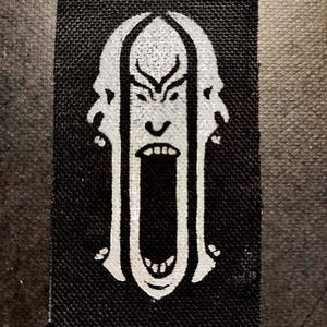 Puede incluir: Un parche rectangular negro con un diseño de rostro blanco estilizado con la boca abierta y gritando. El diseño incluye líneas verticales que sugieren una jaula alrededor de la cara. El parche tiene una superficie texturizada.