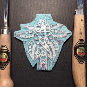 May include: A cyan and white carved linoleum block with a detailed design, flanked by two wood-handled carving tools. The tools have metal blades and labels with cherries. The linoleum block has a detailed, symmetrical design.