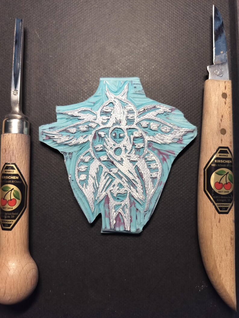May include: A cyan and white carved linoleum block with a detailed design, flanked by two wood-handled carving tools. The tools have metal blades and labels with cherries. The linoleum block has a detailed, symmetrical design.