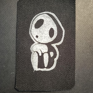 May include: Black rectangular patch with a white cartoon ghost-like figure. The figure has large oval eyes and a small mouth. The patch is made of a textured fabric.
