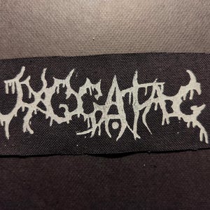 Puede incluir: Un parche rectangular negro con la palabra "EXCAPIATE" en blanco, con una fuente estilizada y goteante. El parche es para ropa o accesorios, con una estética asociada al metal o al punk.