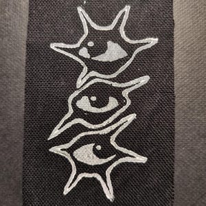 Puede incluir: Parche rectangular negro con un diseño plateado de ojo estilizado. El diseño se compone de tres formas de ojo apiladas con bordes puntiagudos. El parche tiene una superficie texturizada.