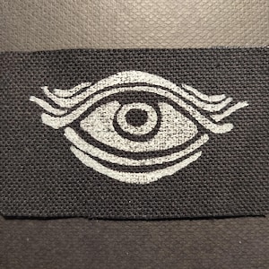 Może przedstawiać: Prostokątna naszywka z ciemnoszarym tłem i białym wzorem oka. Oko jest stylizowane za pomocą zakrzywionych linii powyżej i poniżej oraz okrągłej źrenicy. Naszywka ma teksturowany wygląd.