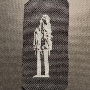 May include: A rectangular black patch featuring a white, stylized figure. The figure resembles a person with long hair or a cloak, standing with legs apart. The patch has a textured surface.