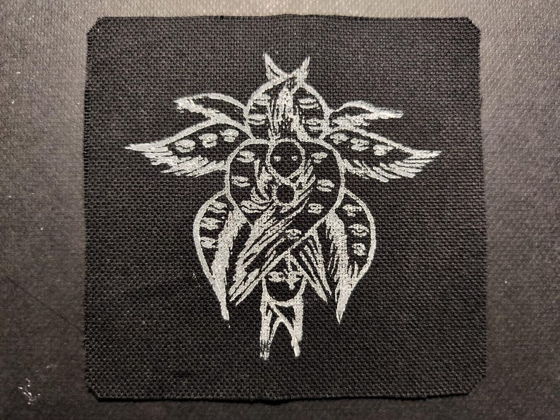 May include: Square black patch with a detailed white illustration of a stylized winged figure. The design features symmetrical elements, including wings, a central heart-like shape, and intricate details.