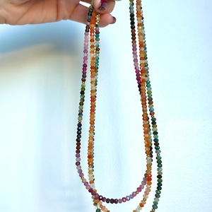 May include: Three strands of colorful beaded necklaces. The beads are small and faceted, with a variety of colors including green, orange, pink, and purple. The necklaces are held up against a white background.