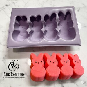 May include: A lavender silicone mould with four bunny-shaped cavities and four pink bunny-shaped soaps. The soaps have black dot eyes. The text "Elite Essentials" is below the mould.