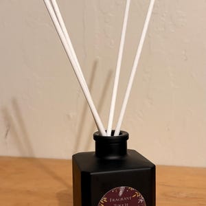 May include: A black square essential oil diffuser with white reeds. The diffuser has a gold-rimmed cork stopper next to it. The label reads "Fragrant Touch" and "Hibiscus Palm".